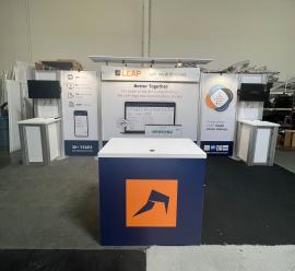 RENTAL: RE-2031 10&rsquo; x 20&rsquo; Inline Design with Extrusion Backwall, Flat Canopy with Black Fabric Cover, (2) RE-1219 Workstation Counters with White Sintra Infill Panels and White Laminated Tops, (1) RE-1202 Reception Counter with Cladded Direct Print Si