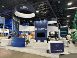 RENTAL: Modified RE-9097 Island Design with 16&rsquo; High Gravitee System Storage Tower, (2) 16 ft. High Curved Walls, (2) Curved Bridged Headers, (3) RE-1575 White Laminated Workstation Counters, (4) RE-1576 White Laminated Counters, (2) RE-703 Charging Sta