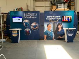 RENTAL: RE-2139 Design with 220" Wide x 90" High Backwall, (2) Monitor Mounts, (4) LED Arm Lights, RE-1201 Tapered Reception Counter, SEG Fabric Graphic, Direct Print Sintra Accent Wing Graphics, and Direct Print Sintra Reception Counter Graphic