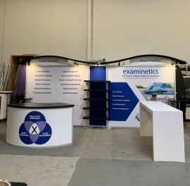 RENTAL: Modified RE-2097 Inline Design with Backwall Frame with Wave Canopies with Black Pillowcase Fabric Covers, (5) LED Arm Lights, RE-1205 Large Curved Reception Counter, (2) Connected Workstation Counters, (5) Black Laminated Shelves, RE-1579 Genius